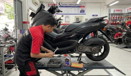 Mekanik berseragam merah-hitam sedang menyervis bagian CVT motor Yamaha NMAX hitam di atas lift hidrolik bengkel resmi.